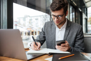 Empresario Sorridente Em Oculos Sentado Junto A Mesa No Cafe Com O Computador Portatil Enquanto Estiver Usando O Smartphone E Escrevendo Algo - ContReal